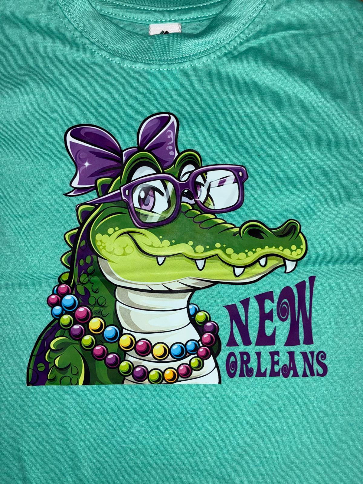 A close-up of a teal heather t-shirt featuring a colorful graphic of a green alligator wearing purple glasses and a purple bow. The text 'NEW ORLEANS' is printed in purple on the left side of the graphic.