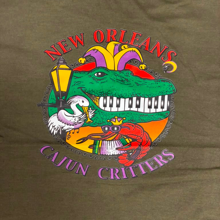 An olive green t-shirt featuring a colorful graphic print of a crocodile playing a piano, with a red alligator on the left and a purple and yellow crown on the right. The text 'CAJUN CRITTERS' is written in purple on the left, and 'NEW ORLEANS' is written in red on the right.