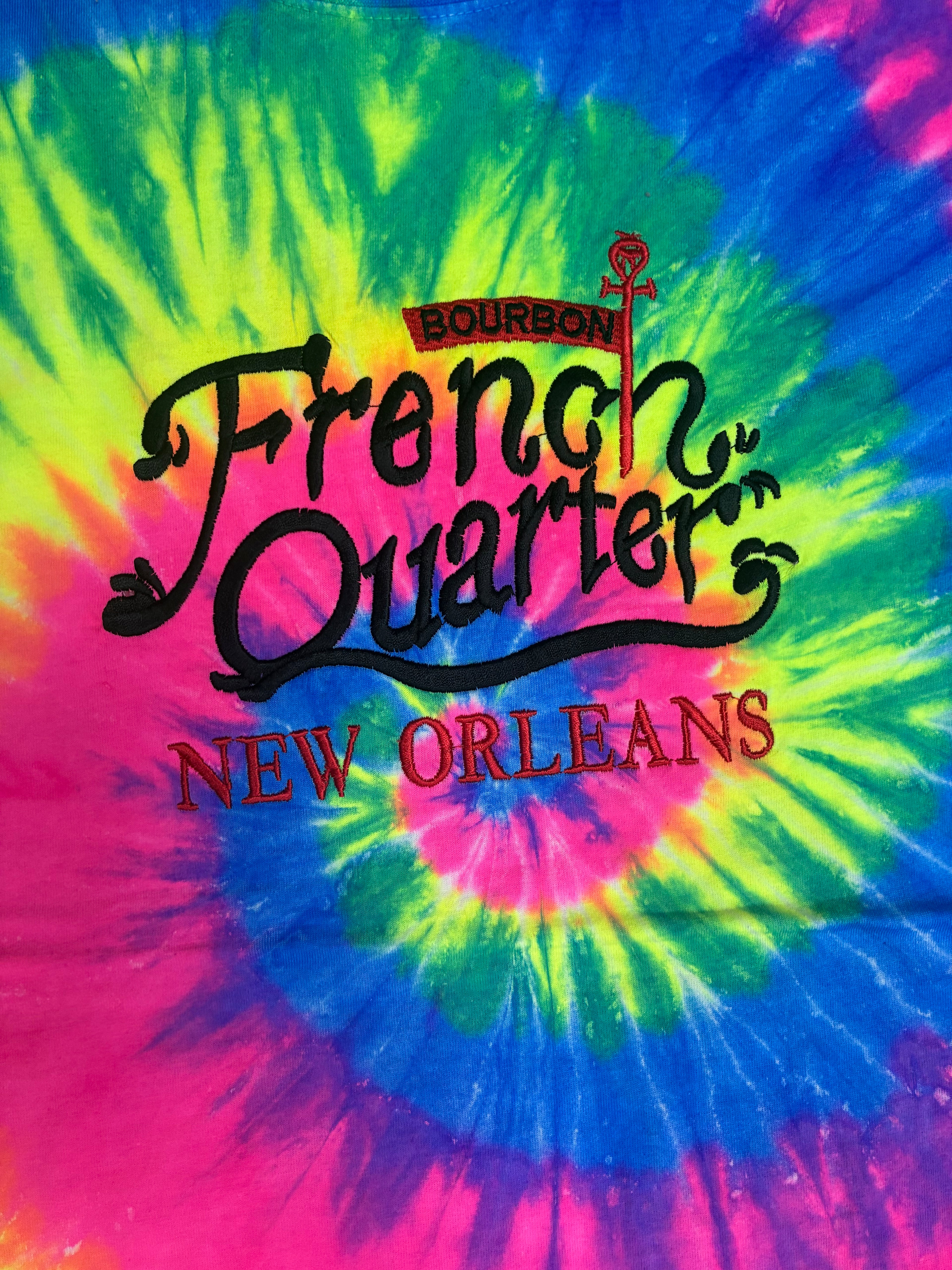 A close-up of a vibrant tie-dye shirt featuring a spiral pattern of neon pink, green, blue, and yellow. The front is printed with the text 'French Quarter New Orleans' in black and red, with a small Bourbon logo above.