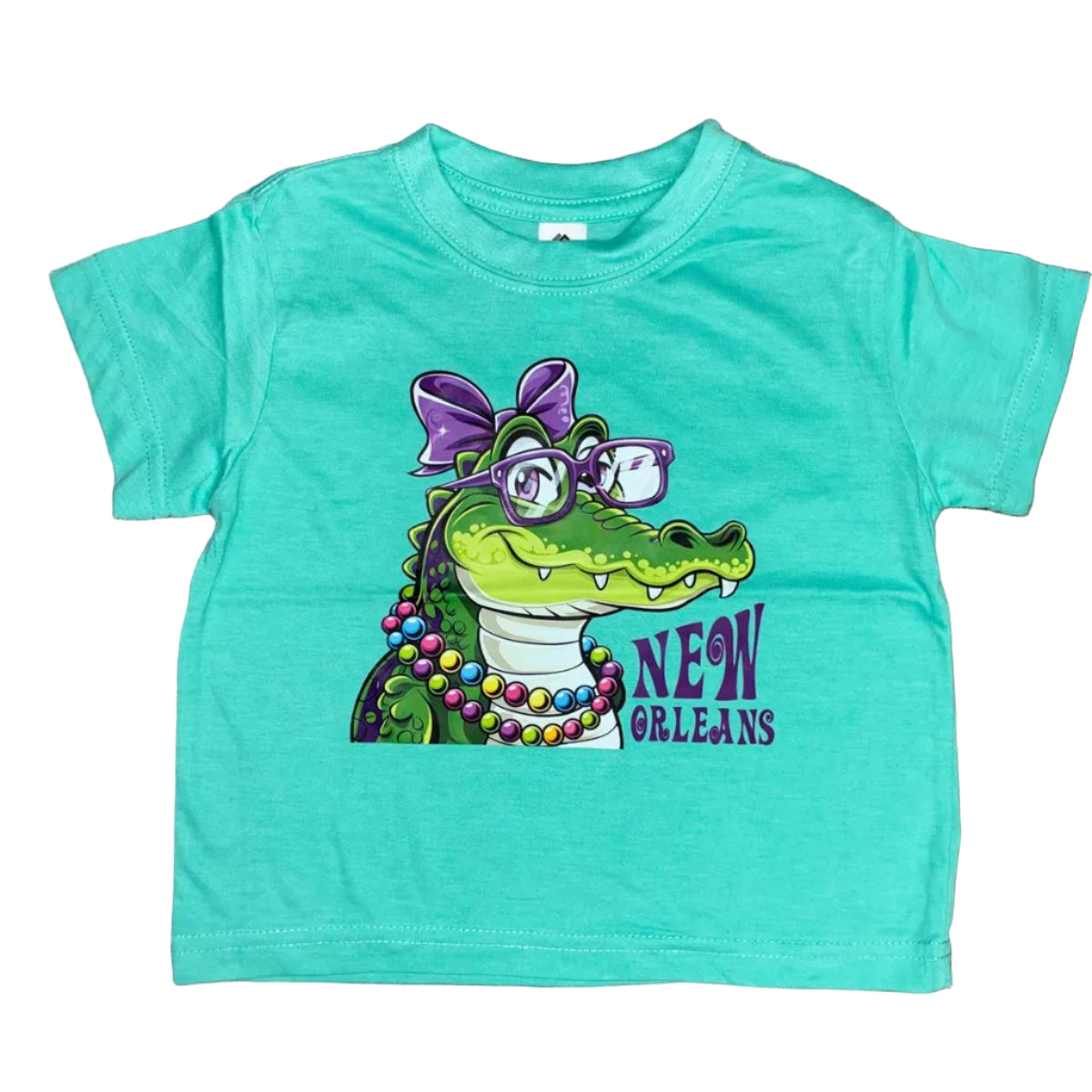 A turquoise short-sleeve toddler t-shirt laid flat on a white background, featuring a colorful graphic of an alligator wearing a bow and beads on the neck with 'New Orleans' text