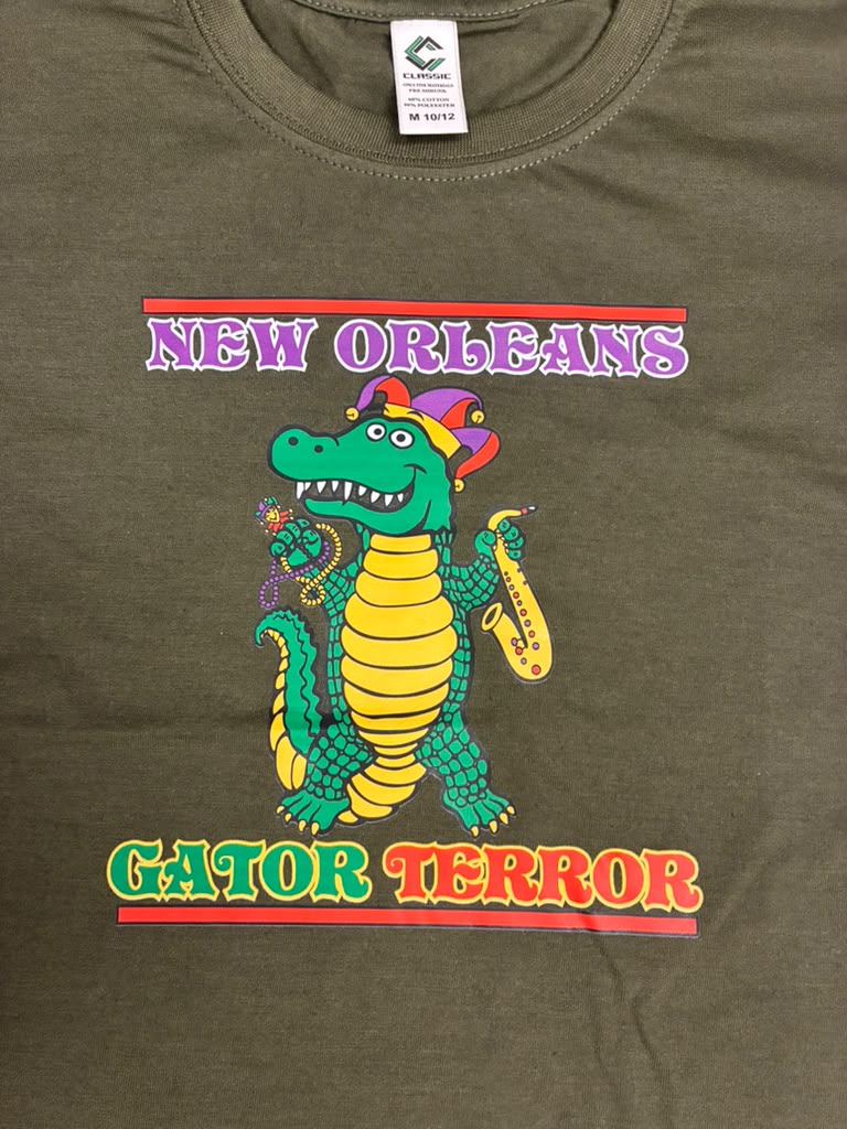 An olive green t-shirt featuring a colorful graphic of a green and yellow alligator wearing a purple and orange hat and playing a saxophone, with 'NEW ORLEANS' above and 'GATOR TERROR' below in stylized text.