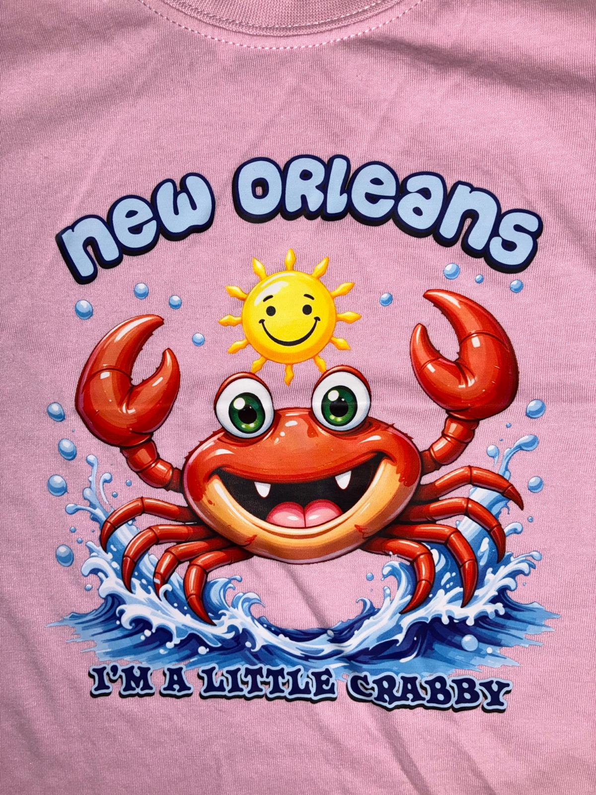 A close-up of a pink graphic t-shirt featuring a cartoon red crab, a yellow smiling sun, and a crab on blue waves with the text 'NEW ORLEANS' and 'I'M A LITTLE CRABBY'.