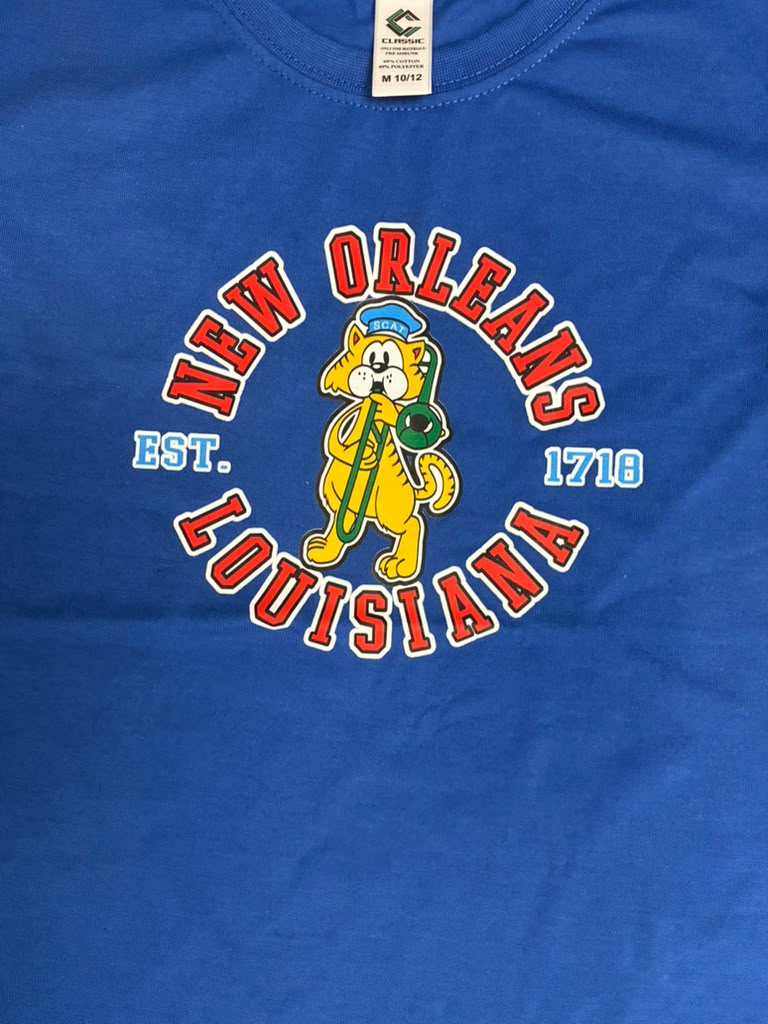 A close-up of a royal blue t-shirt featuring a vintage-style graphic of the New Orleans mascot, a yellow dog playing a green saxophone, surrounded by the text 'NEW ORLEANS LOUISIANA' in red and white, with 'EST. 1718' on either side.