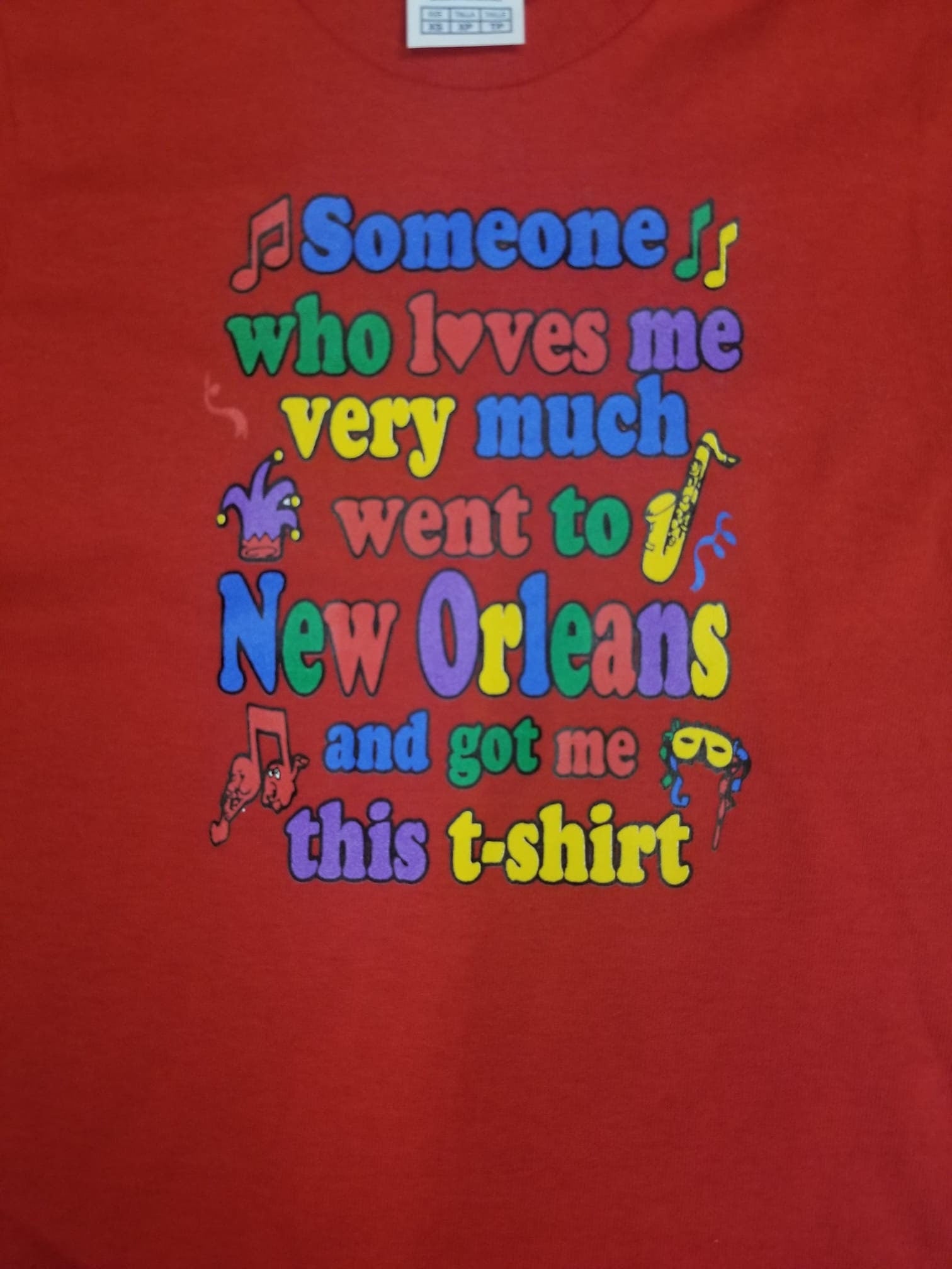 A red short-sleeve t-shirt featuring a colorful retro-style graphic that reads 'Someone who loves me very much went to New Orleans and got me this t-shirt' in multicolored letters with musical notes and a jester illustration.