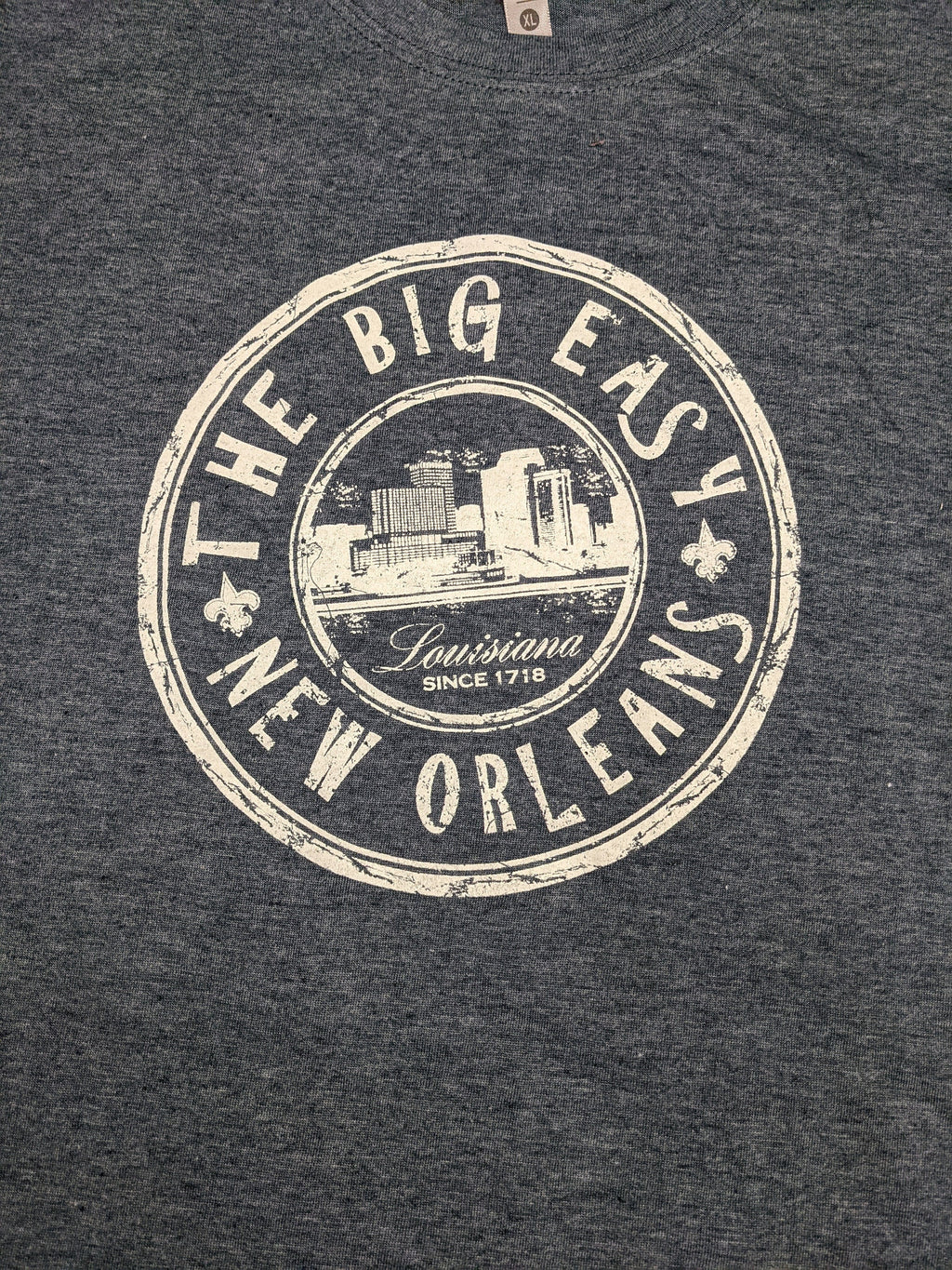 Close-up of a dark heather gray t-shirt featuring a distressed cream-colored circular graphic that reads 'THE BIG EASY NEW ORLEANS' with a cityscape illustration and 'Louisiana Since 1718' in the center.