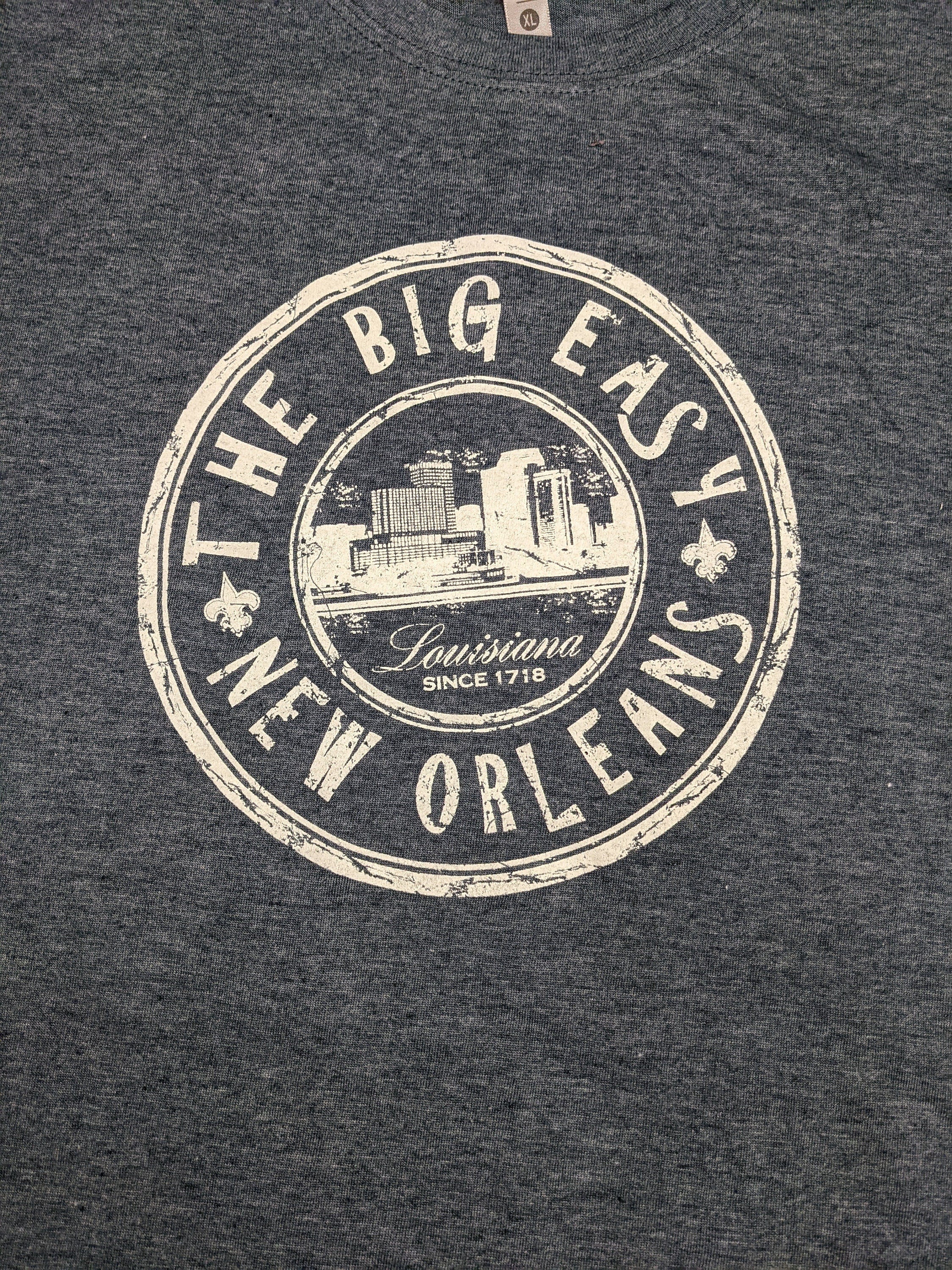 Close-up of a dark heather gray t-shirt featuring a distressed cream-colored circular graphic that reads 'THE BIG EASY NEW ORLEANS' with a cityscape illustration and 'Louisiana Since 1718' in the center.