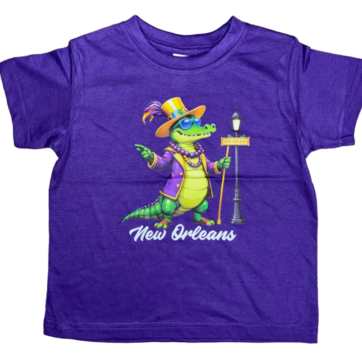 A purple short-sleeve kids' t-shirt laid flat on a wooden surface, featuring a colorful graphic of a Mardi Gras alligator wearing a top hat and beads, with the text 'New Orleans' printed below.