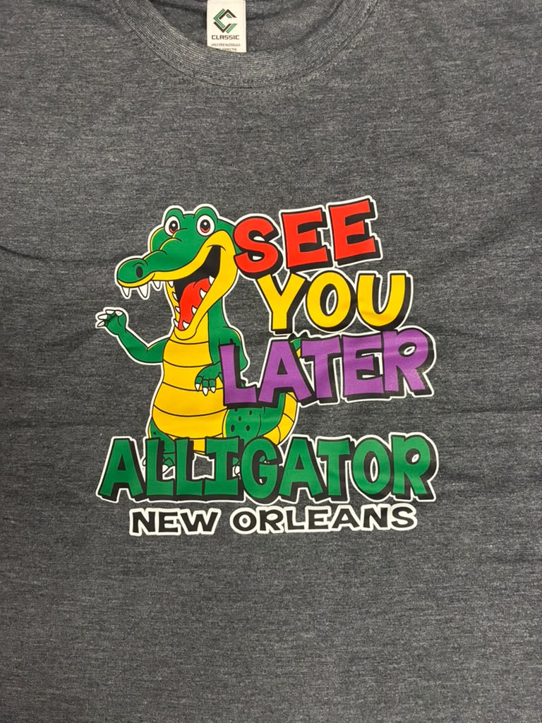 A close-up of a heather gray t-shirt featuring a colorful graphic of a cartoon alligator next to the text 'SEE YOU LATER ALLIGATOR NEW ORLEANS' in bold, multi-colored letters.