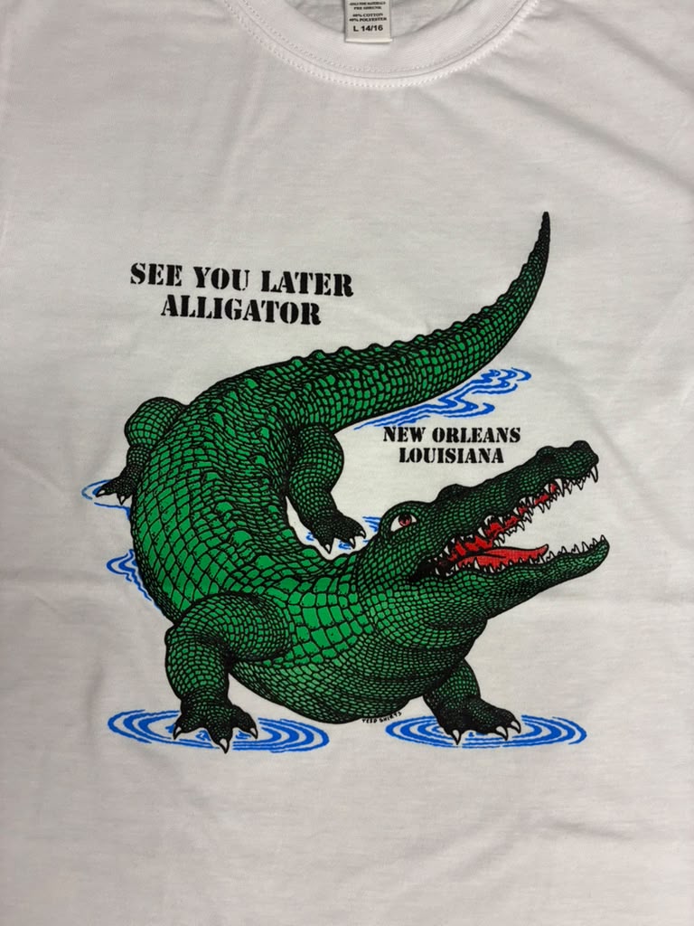 A white short-sleeve t-shirt featuring a graphic print of a green alligator in water with blue ripples, accompanied by the text 'SEE YOU LATER ALLIGATOR' and 'NEW ORLEANS LOUISIANA'.