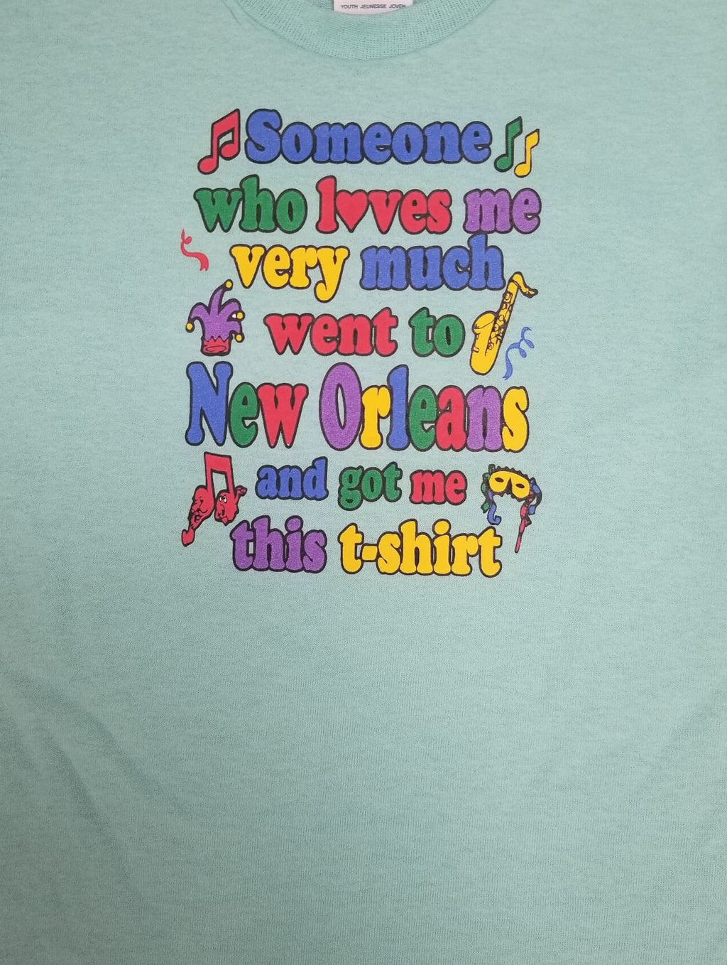 A close-up of a light mint green t-shirt featuring a colorful graphic print with the text 'Someone who loves me very much went to New Orleans and got me this t-shirt' in a playful, multi-colored font with small jazz-themed illustrations.