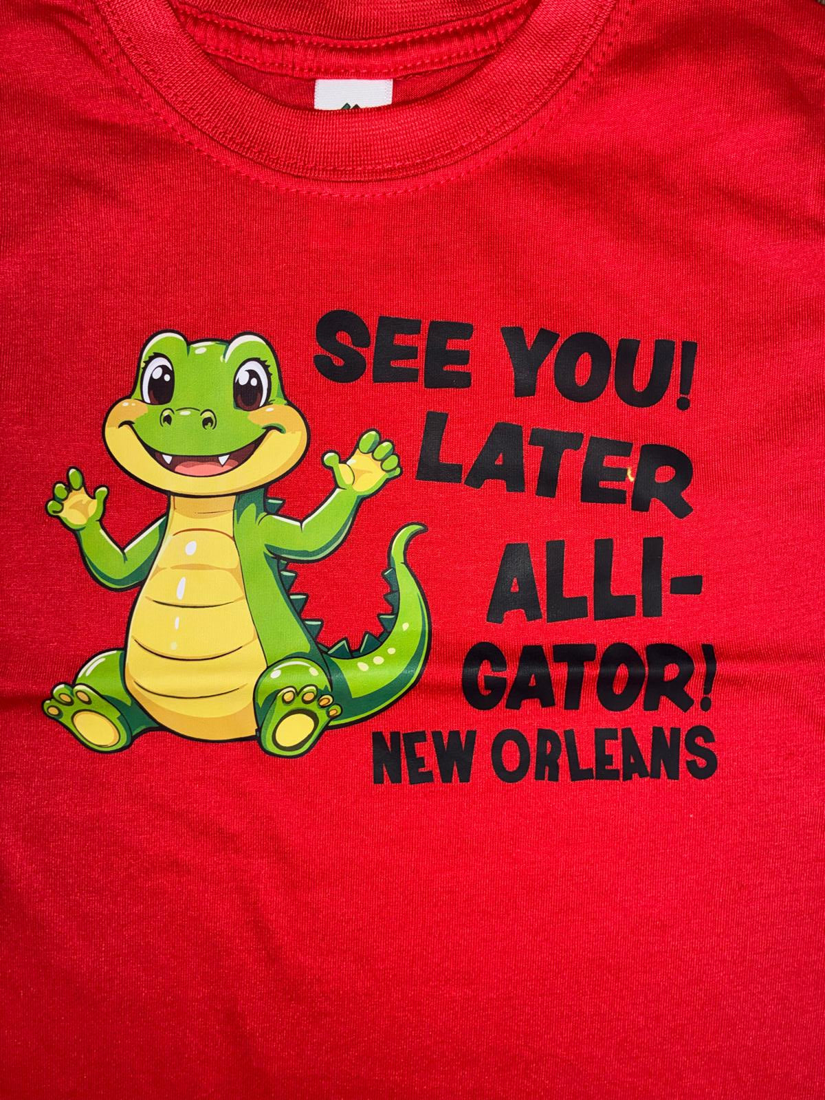 A close-up of a red t-shirt featuring a cartoon green alligator graphic and the text 'SEE YOU! ALLI- GATOR! NEW ORLEANS' in black lettering.