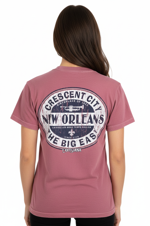 A woman viewed from the back wearing a dusty rose short-sleeve t-shirt with a large distressed circular graphic on the back featuring the text 'Crescent City New Orleans The Big Easy' and a fleur-de-lis.