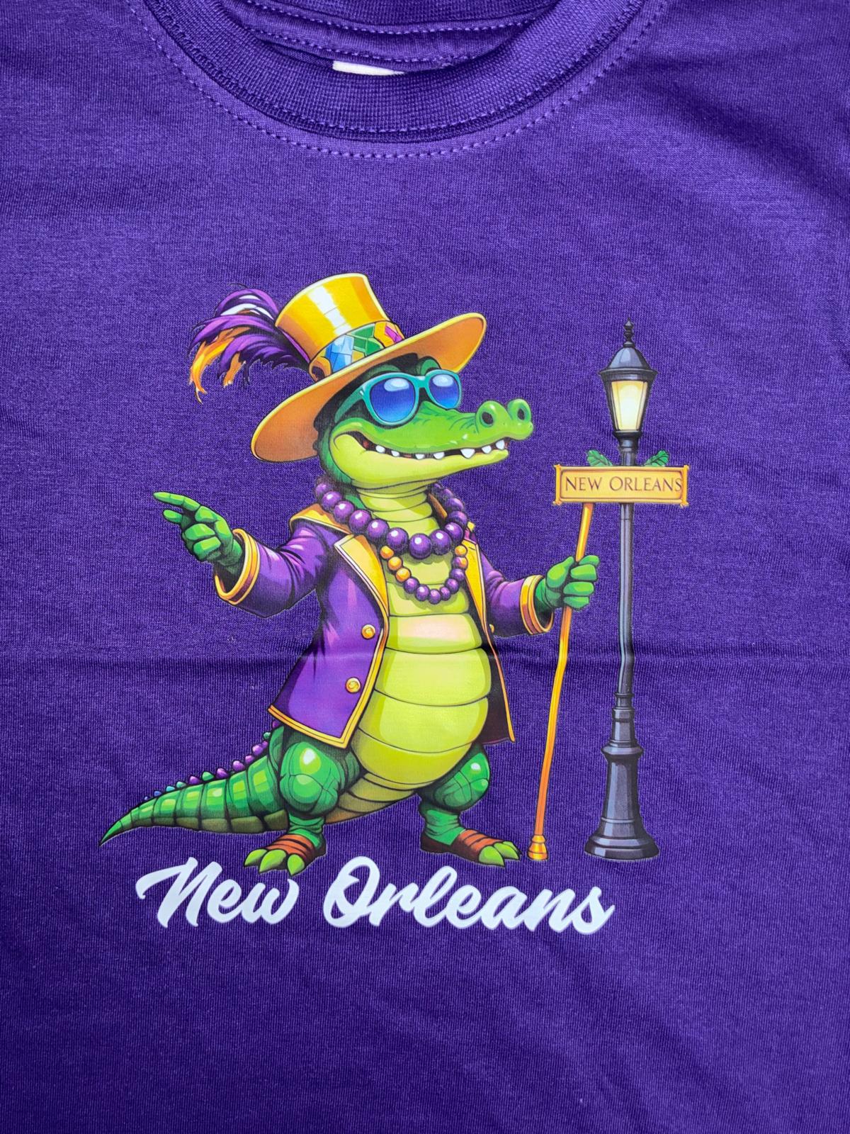 A close-up of a purple t-shirt featuring a colorful graphic of a cartoon alligator dressed in Mardi Gras attire with a New Orleans street sign and the words 'New Orleans' printed below.
