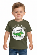 A toddler boy wearing an olive green t-shirt with a circular graphic that reads 'NEW ORLEANS IT'S GREAT TO BE A CATOR!' around a cartoon green alligator, paired with blue jeans, on a solid white background.