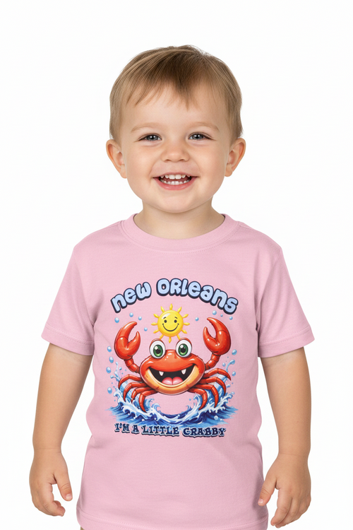 A smiling toddler wearing a light pink short-sleeve t-shirt with a colorful cartoon crab graphic and the text 'new orleans' and 'i'm a little crappy' on a plain white background.
