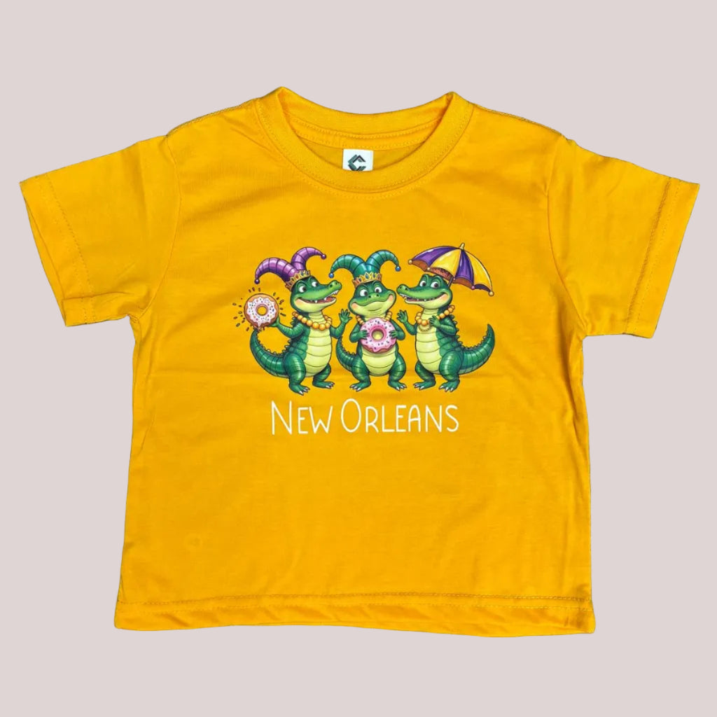 A toddler yellow short-sleeve t-shirt with a colorful graphic of three green alligators holding donuts and an umbrella, with the text 'New Orleans' printed below. 