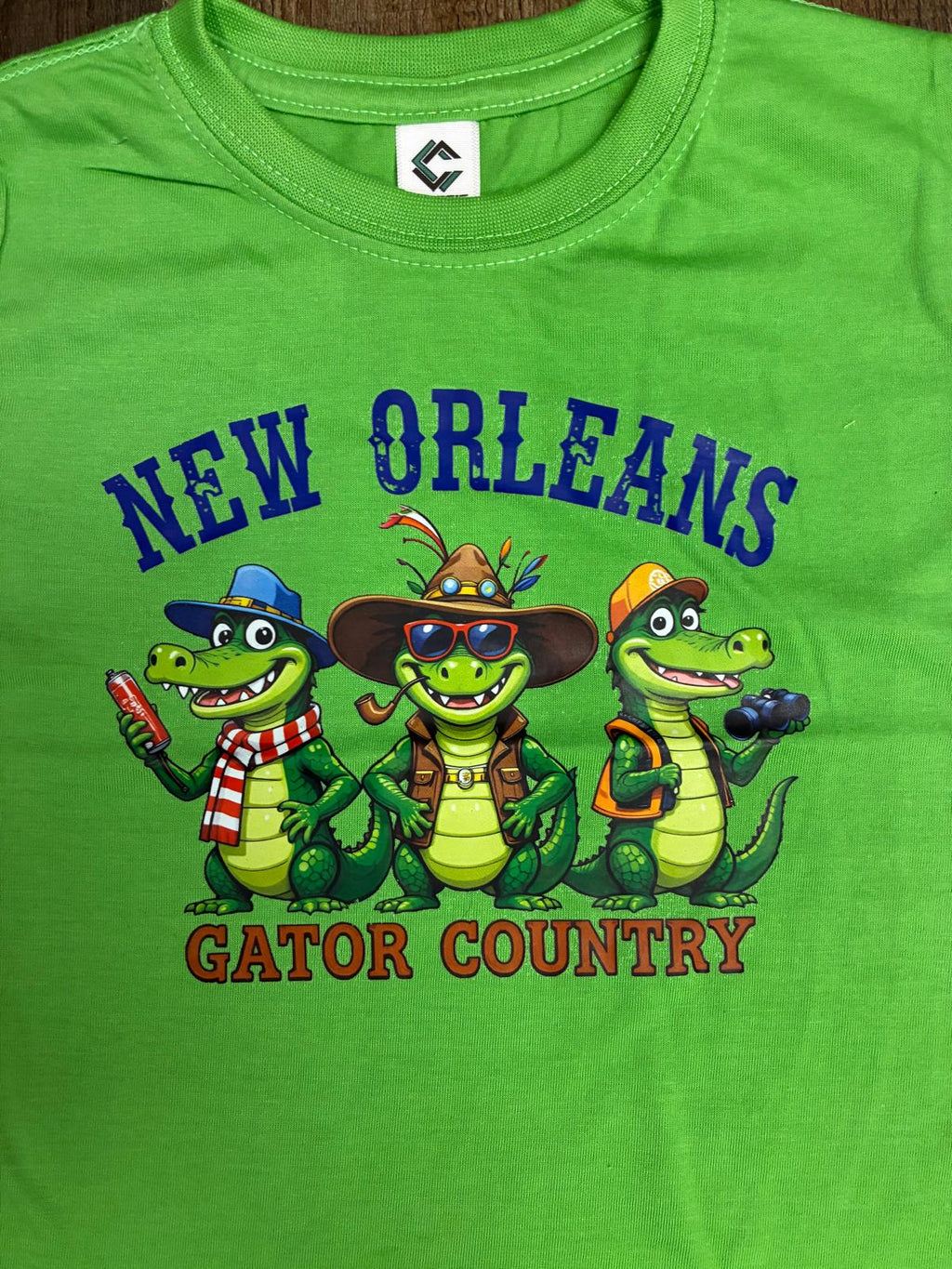 A bright green short-sleeve t-shirt featuring a cartoon graphic of three gators in New Orleans-themed attire with the text 'NEW ORLEANS GATOR COUNTRY'.